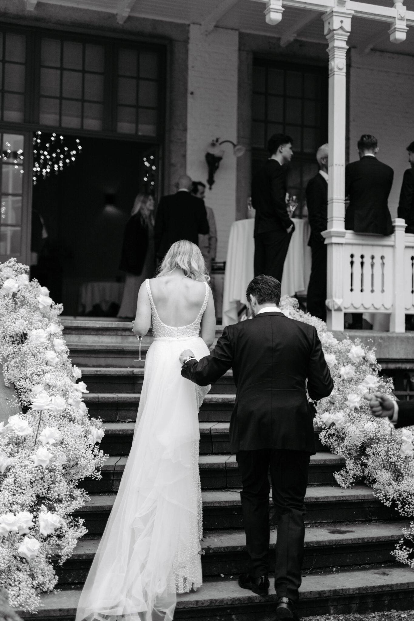 Braut und Bräutigam schreiten die blumengeschmückten Stufen einer atemberaubenden Hochzeit an einem Reiseziel hinauf, umgeben von Gästen in einem wunderschönen Außenbereich.