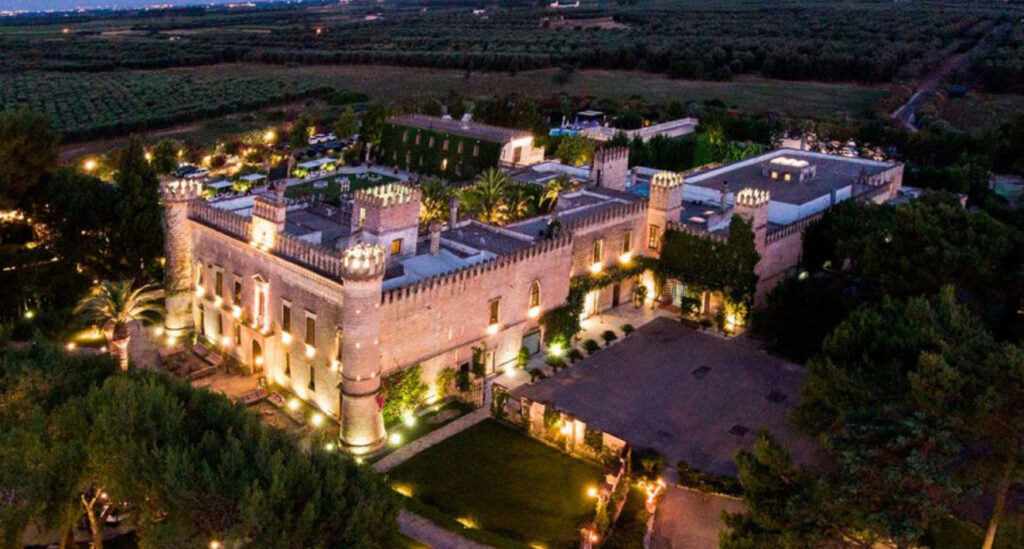 Luftaufnahme eines großen, beleuchteten Schlosses inmitten von Grün und Ackerland in der Abenddämmerung - eine zauberhafte Kulisse, die sich perfekt für eine Hochzeit am Reiseziel oder für diejenigen eignet, die vom Heiraten in Italien träumen.