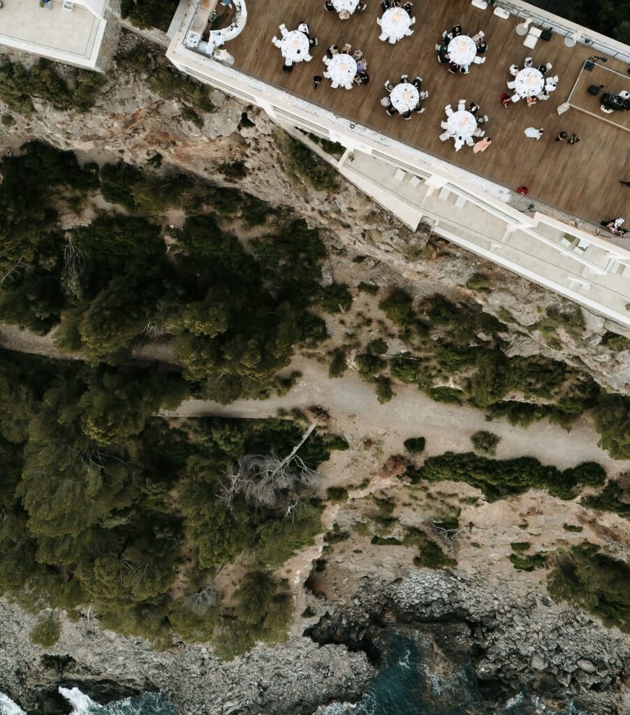 Luftaufnahme einer Veranstaltung im Freien mit runden Tischen in der Nähe einer bewaldeten Klippe oberhalb der felsigen Küste – ein atemberaubender Ort auf Mallorca, der jeden Hochzeitsplaner begeistern würde.