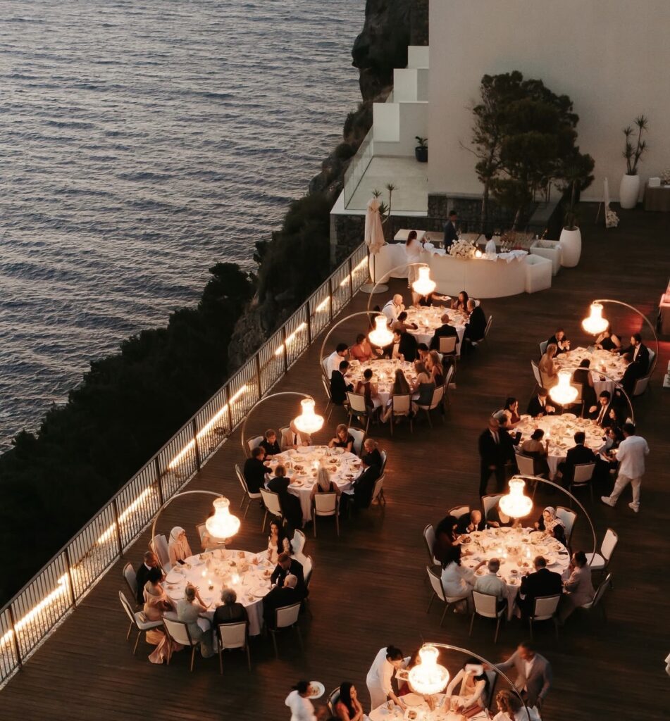 Eine Abendveranstaltung im Freien mit runden Tischen, Gästen beim Essen und Meerblick auf einer Terrasse direkt am Wasser – die perfekte Hochzeitslocation auf Mallorca für eine unvergessliche Feier.