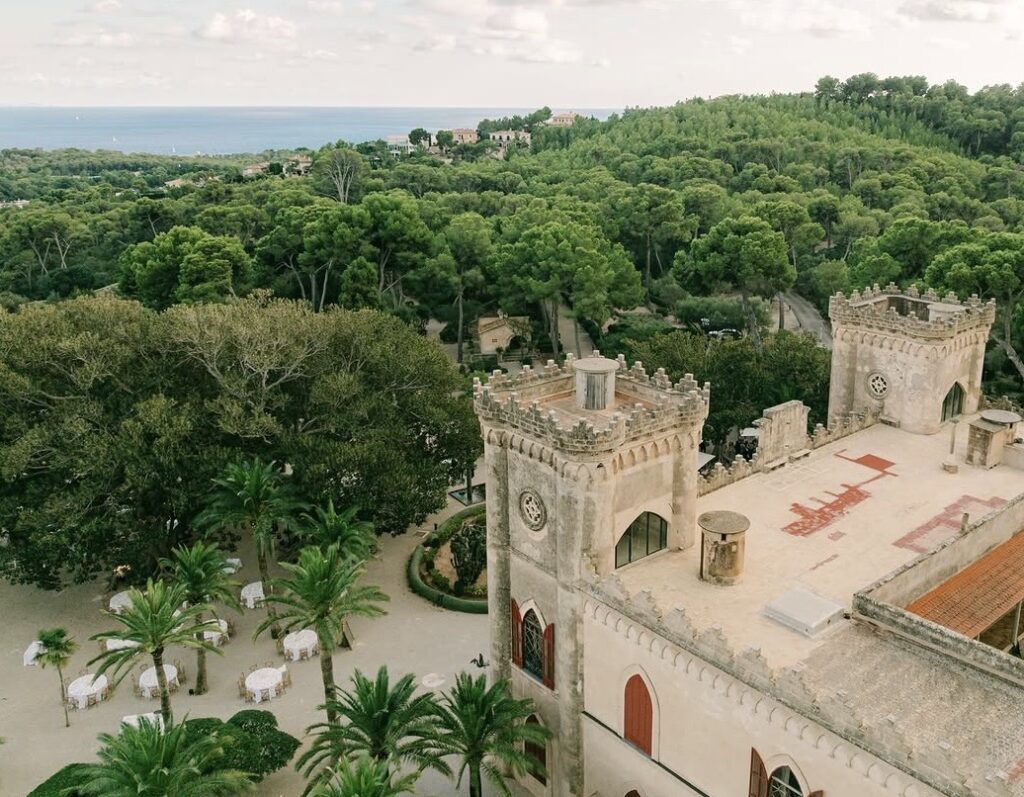 Ein Steinschloss mit zwei Türmen, umgeben von Bäumen und Palmen, mit Blick auf das Meer im Hintergrund – eine idyllische Hochzeitslocation auf Mallorca, perfekt für die Feier Ihres besonderen Tages. Lassen Sie sich von einem erfahrenen Hochzeitsplaner dabei unterstützen, in dieser romantischen Kulisse unvergessliche Erinnerungen zu schaffen.
