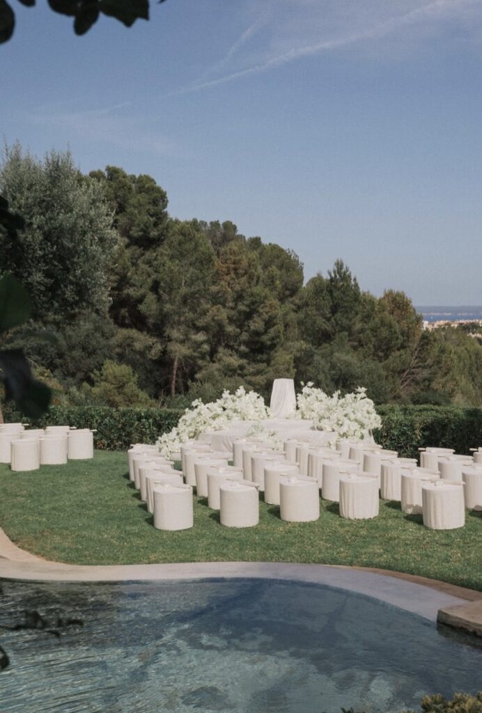 Eine Hochzeitskulisse im Freien mit weißen Stühlen, einem mit Blumen geschmückten Altar und einem Pool, umrahmt von Bäumen und einem strahlend blauen Himmel – ein idealer Ort auf Mallorca für Ihre Traumhochzeit. Lassen Sie einen erfahrenen Hochzeitsplaner dafür sorgen, dass jedes Detail perfekt ist.