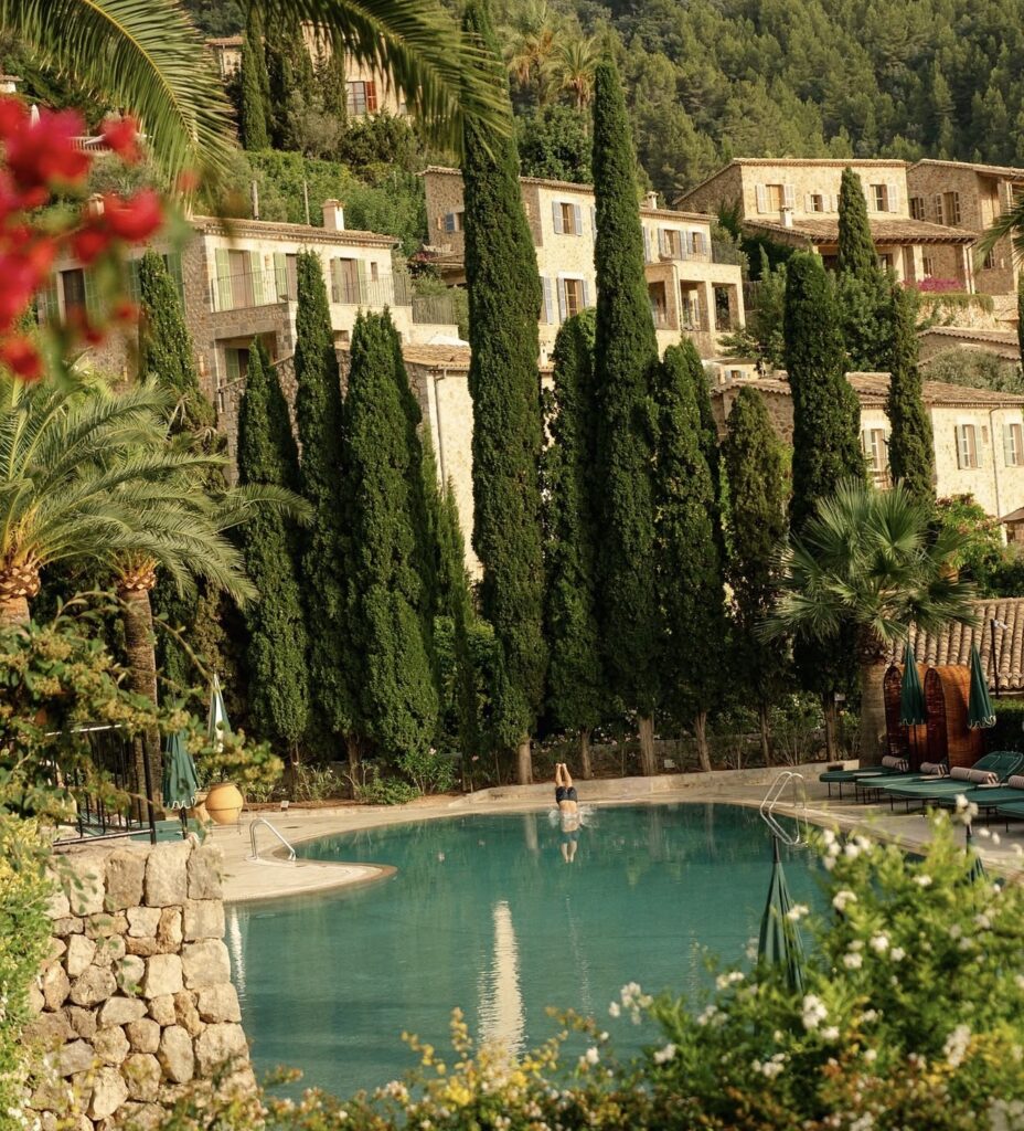 Eine Person steht in einem Pool auf Mallorca, umgeben von hohen Bäumen und Steinhäusern am Hang, die von üppigem Grün umgeben sind.