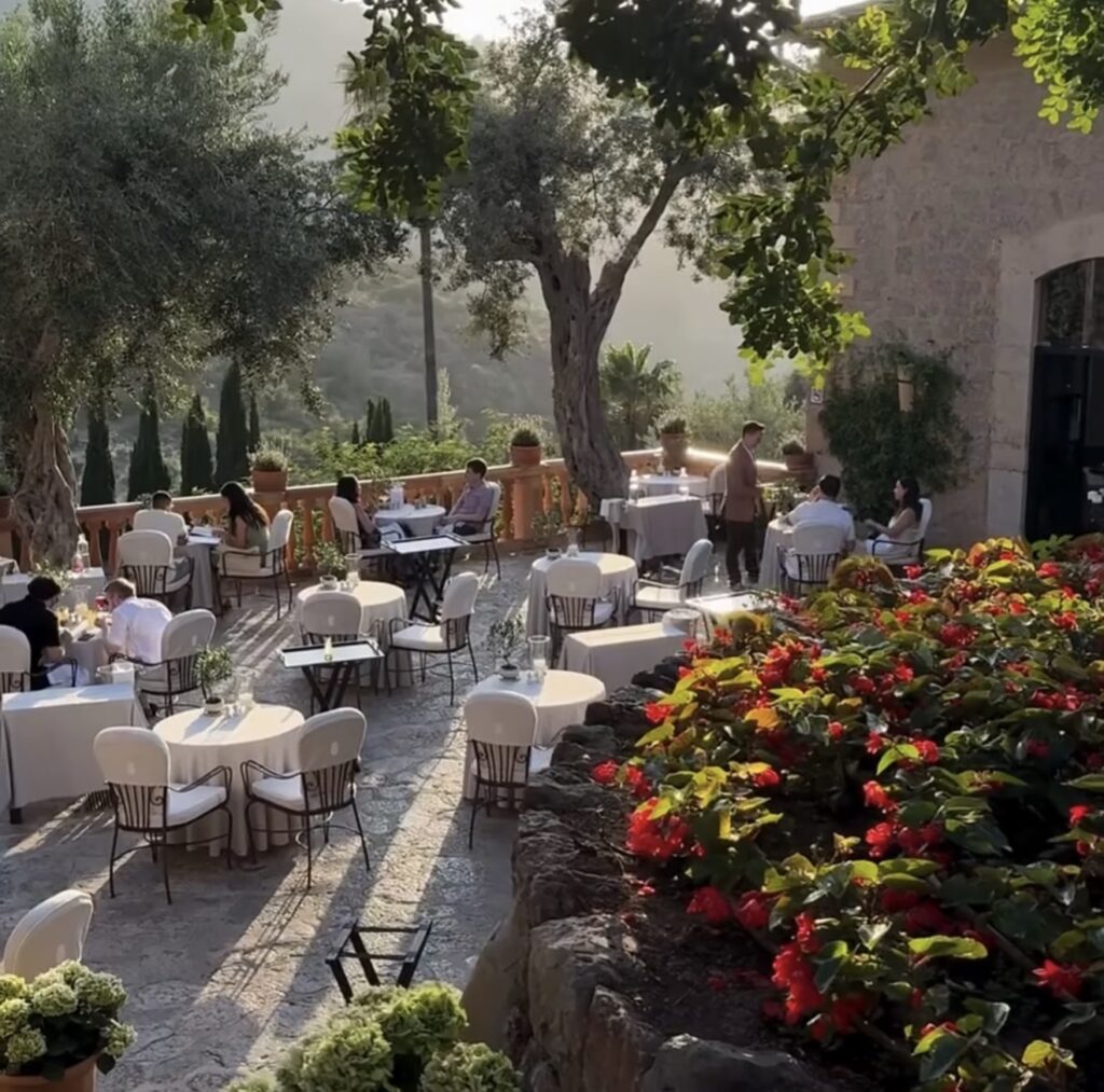 Sonnige Restaurantterrasse im Freien, ideal als Hochzeitslocation, mit weißen Tischen, üppigen Blumen und Steinmauern.