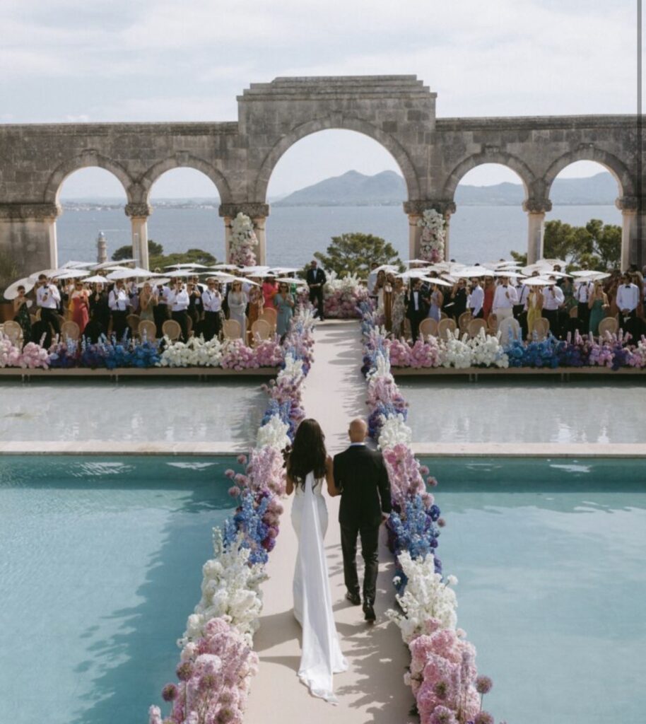 Ein Brautpaar schreitet auf einer Hochzeitslocation auf Mallorca einen mit Blumen geschmückten Gang am Pool entlang, im Hintergrund sind Berge zu sehen.