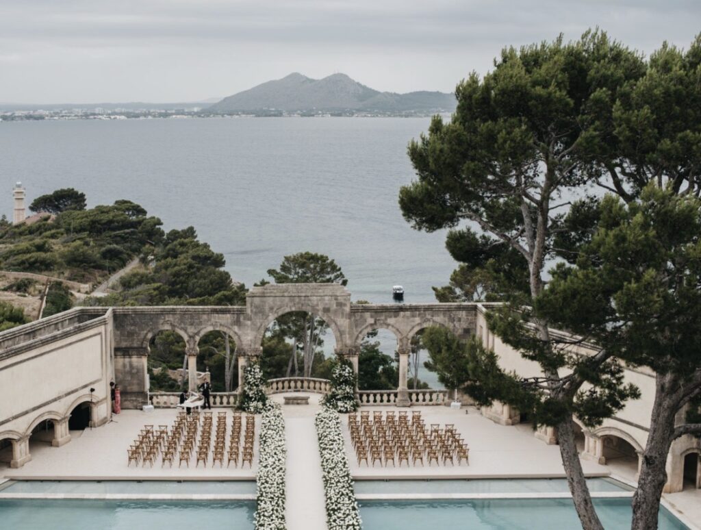 Atemberaubende Hochzeitslocation auf Mallorca direkt am Meer, mit einem mit Blumen geschmückten Gang, Stuhlreihen und Blick auf die Berge.