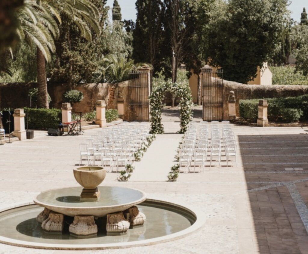 Hochzeitsdekoration im Freien in einem sonnigen Innenhof auf Mallorca mit Stuhlreihen, einem Blumenbogen und einem Springbrunnen.