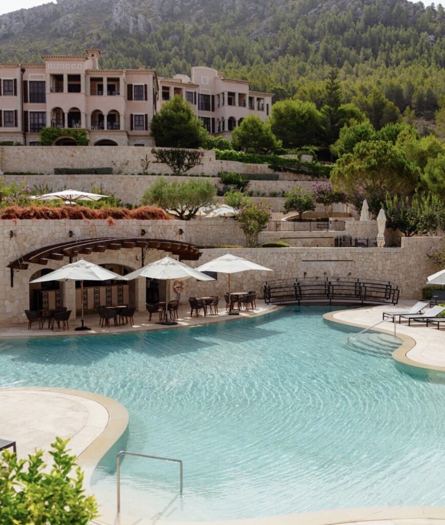 Ein luxuriöses Resort auf Mallorca in Hanglage mit einem großen Pool, Sonnenschirmen und üppig grünen Bäumen im Hintergrund.