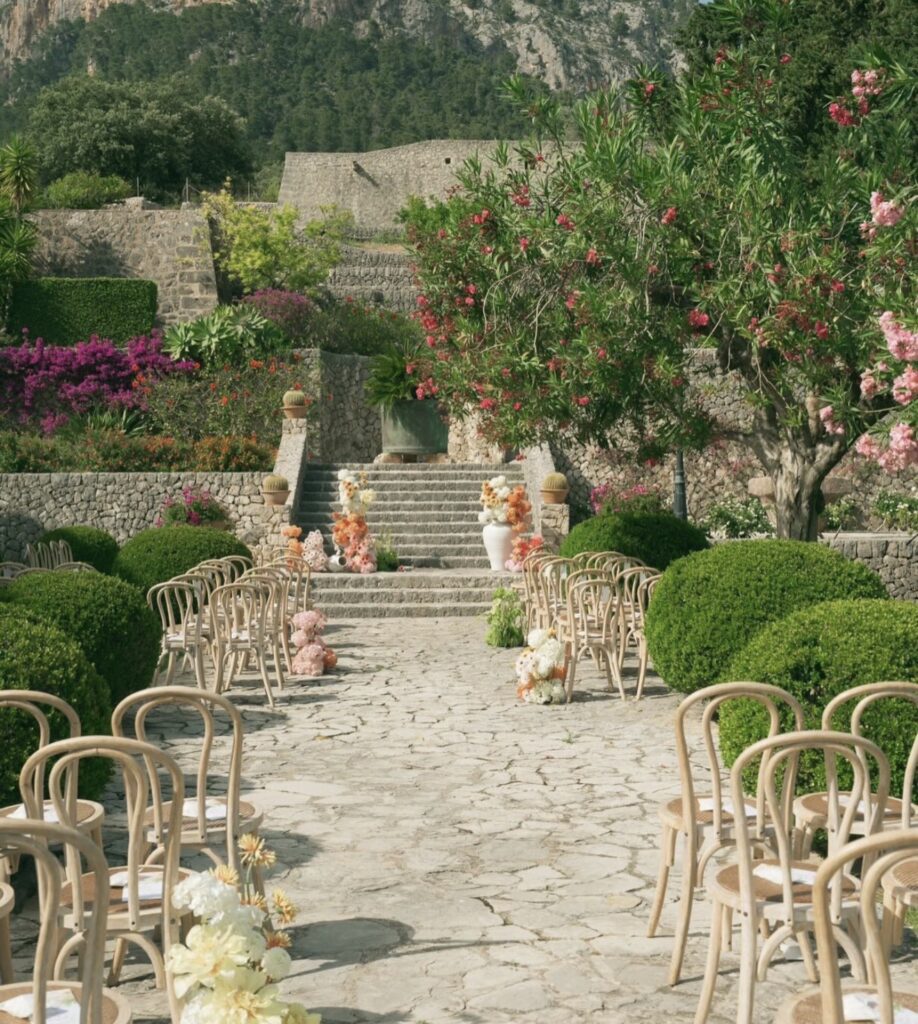 Hochzeitsgang im Freien in einem üppigen mallorquinischen Garten – Holzstühle, Blumen, Steinweg an einer traumhaften Hochzeitslocation.