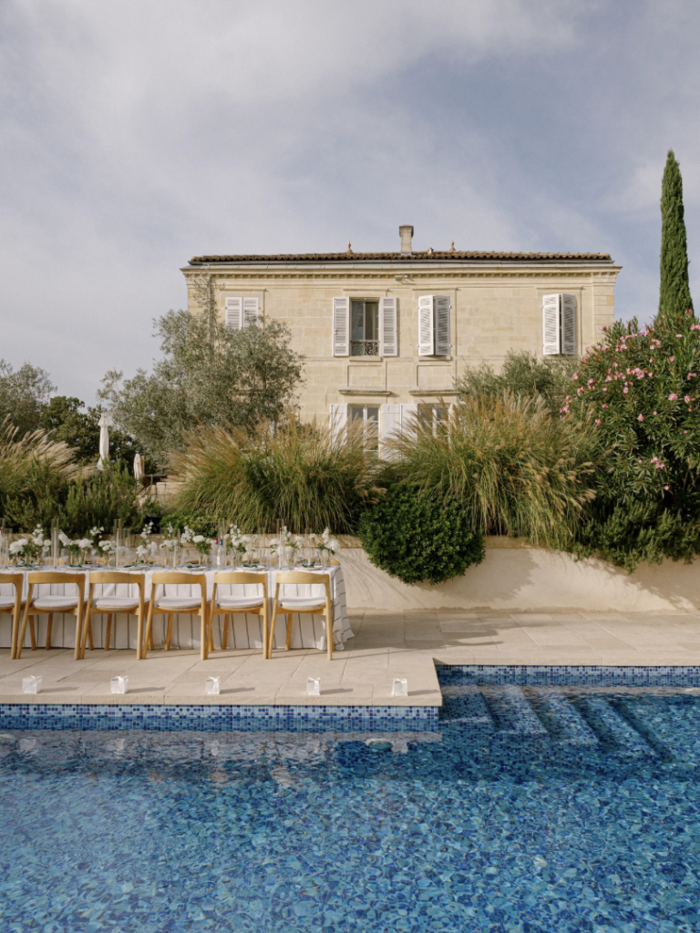 Ein langer Tisch mit weißen Stühlen am blauen Pool, vor einem großen Steinhaus und üppigem Grün - perfekt für ein Heiraten in Italien, das einen idyllischen Rahmen bietet, kuratiert von einem professionellen Hochzeitsplaner Italien.
