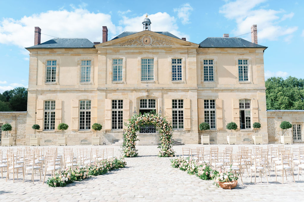Elegante Hochzeit im Freien mit Blumenbogen und Stühlen vor einem großen französischen Schloss unter blauem Himmel, perfekt für alle, die von einer Luxushochzeit in Italien träumen.