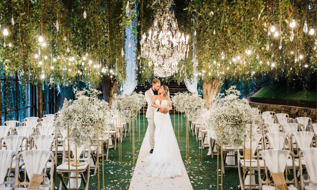 Eine Braut und ein Bräutigam umarmen sich unter Kronleuchtern in einem mit Blumen und Grünzeug geschmückten Hochzeitsgang im Freien.