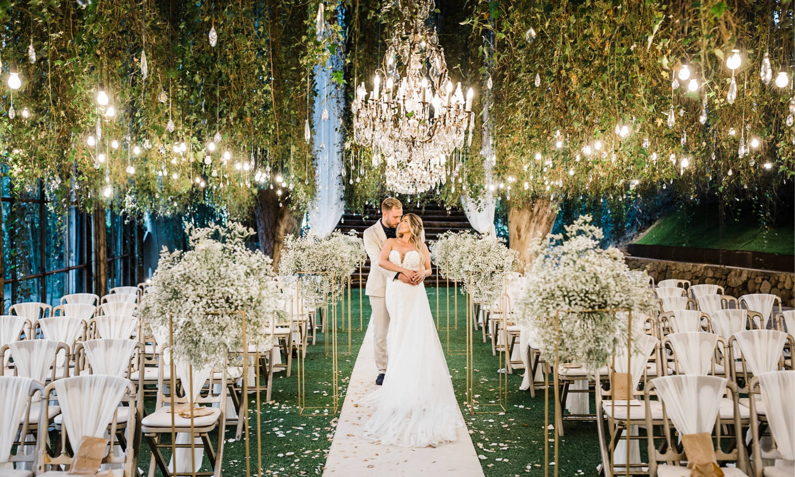 Eine Braut und ein Bräutigam umarmen sich unter Kronleuchtern in einem mit Blumen und Grünzeug geschmückten Hochzeitsgang im Freien.