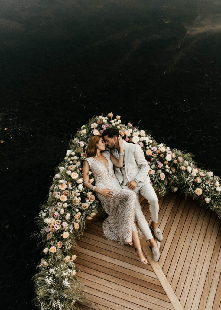 Ein formell gekleidetes Paar liegt auf einem blumengeschmückten Holzboot, umgeben von dunklem Wasser.
