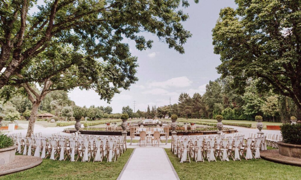 Hochzeitszeremonie im Freien mit weißen Stuhlreihen und einem Mittelgang, umgeben von Bäumen und Grünpflanzen.