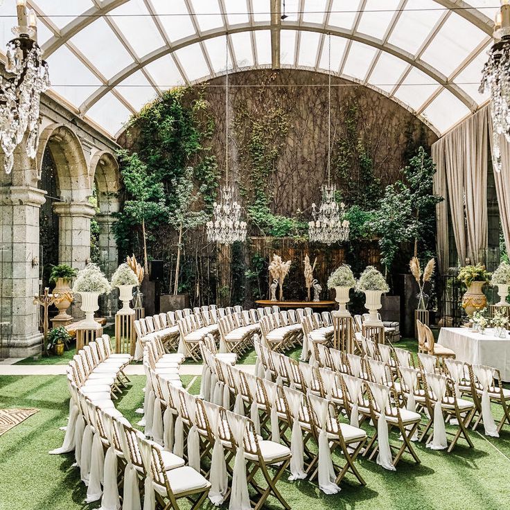 Elegante Hochzeitszeremonie im Innenbereich mit weißen Stühlen, Kronleuchtern, Grünpflanzen und gewölbten Steinwänden.