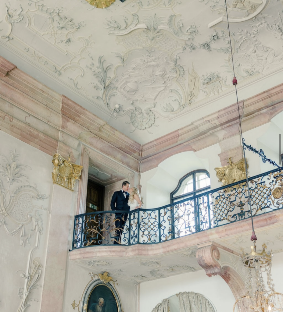 In den eleganten, aufwändig dekorierten Sälen von Schloss Leopoldskron steht ein Paar gemeinsam auf einem kunstvollen Balkon, der mit seinen hohen Decken und der herrschaftlichen Umgebung eine zeitlose Hochzeitsatmosphäre schafft.