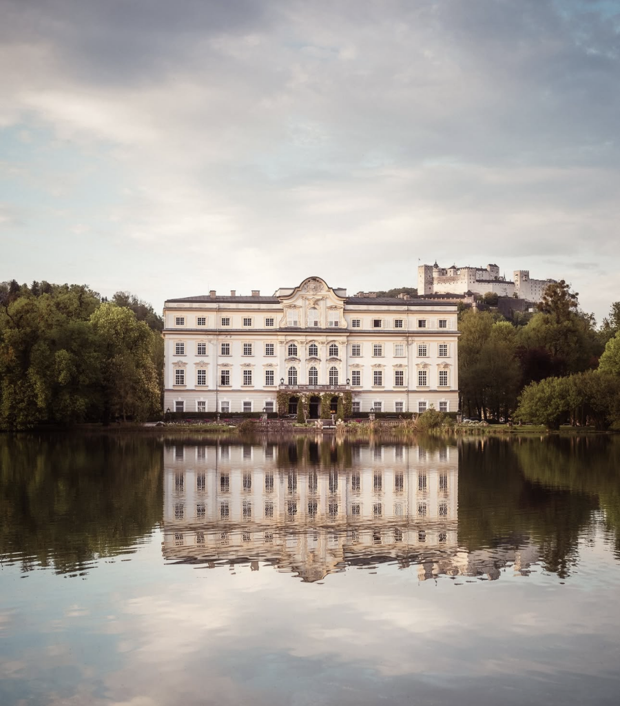Das große weiße Schloss Leopoldskron spiegelt sich in einem ruhigen See, umgeben von Bäumen, mit einem Schloss auf einem Hügel im Hintergrund - eine bezaubernde Kulisse, die perfekt für eine Hochzeitsfeier ist.