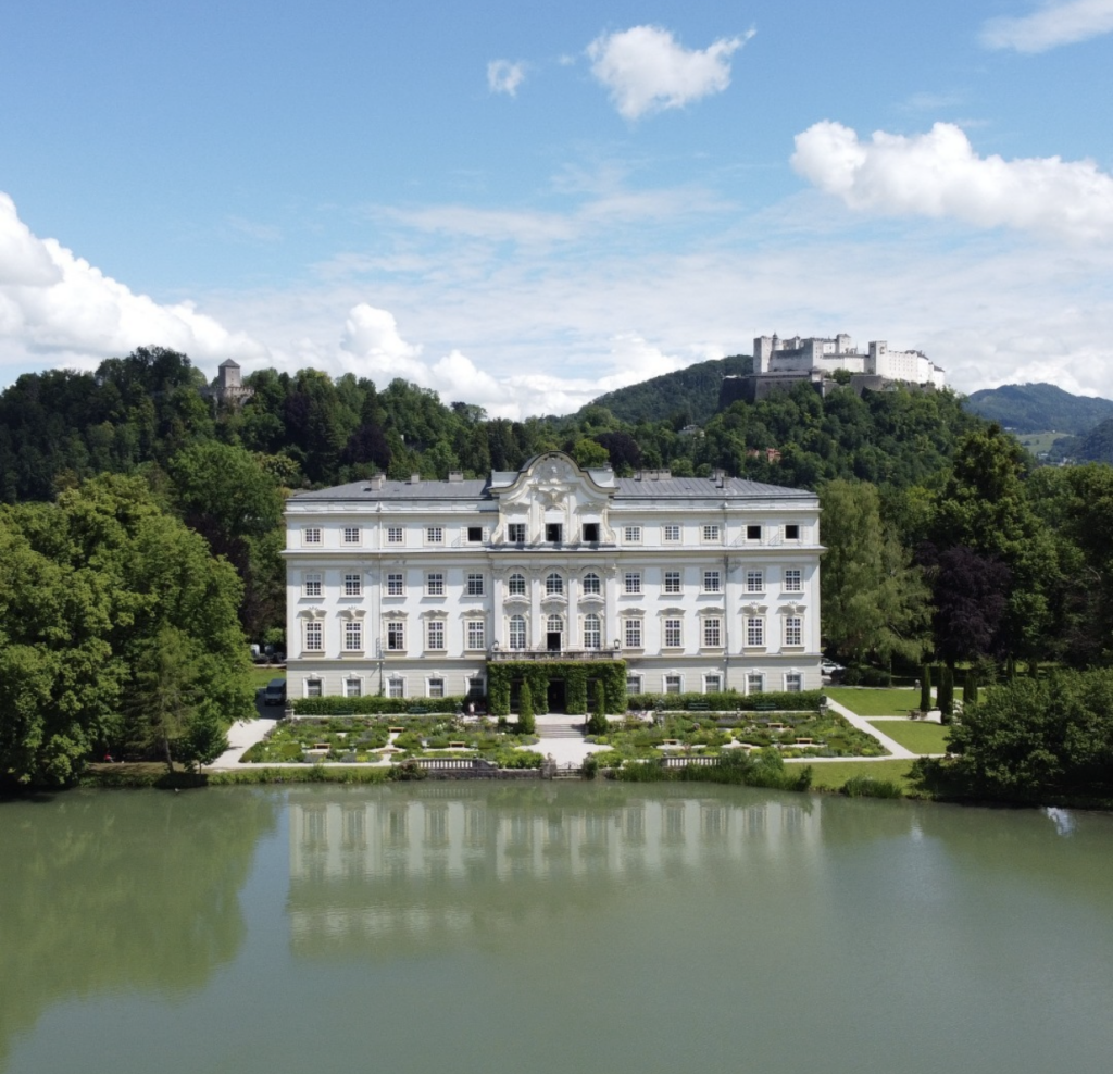 Ein prächtiges weißes Schloss, Schloss Leopoldskron, steht an einem See - ein bezauberndes Hochzeitsschloss mit grünen Hügeln und einer Festung im Hintergrund unter einem blauen Himmel.