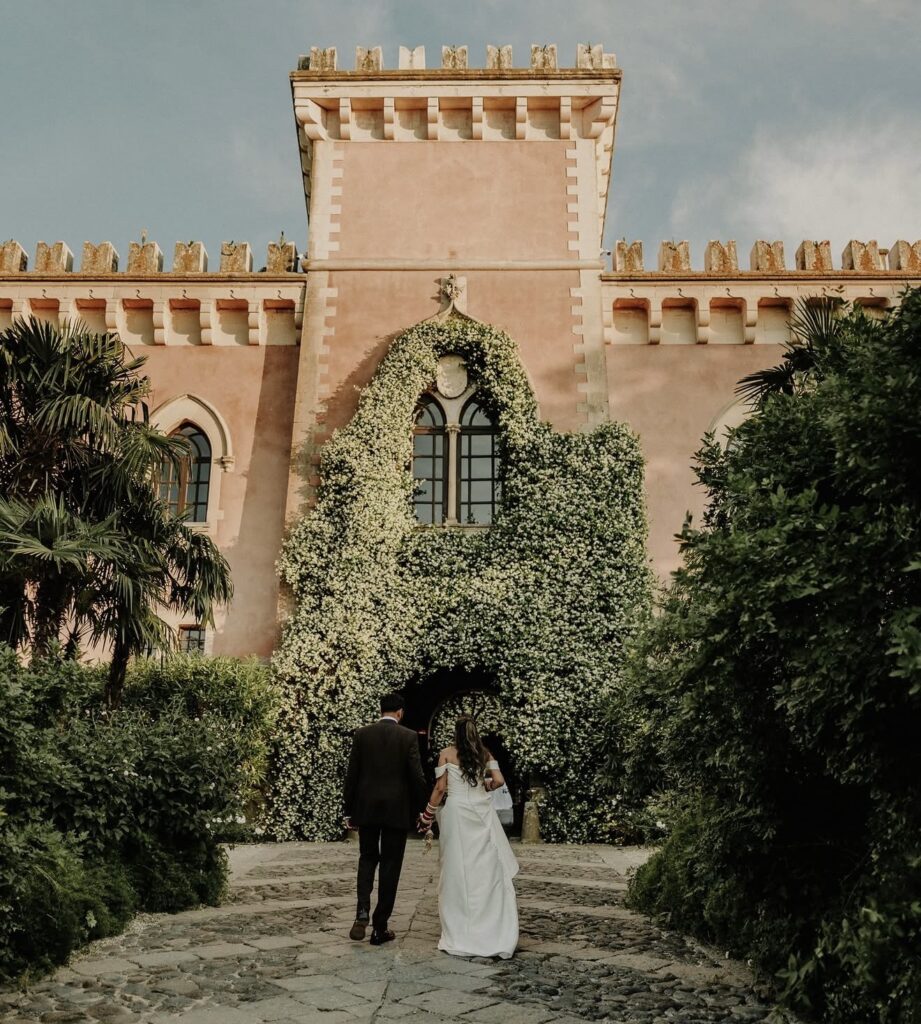 Ein Brautpaar schreitet auf ein prächtiges, mit Efeu bewachsenes Gebäude mit Bogenfenstern und einem Steinweg zu und verkörpert damit die zeitlose Romantik von „Heiraten auf Sizilien“.
