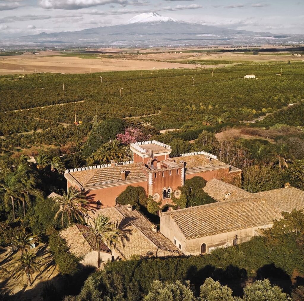 Luftaufnahme einer historischen Villa, umgeben von Feldern und Bäumen, mit einem schneebedeckten Berg im Hintergrund – ein bezaubernder Ort für eine Hochzeit auf Sizilien.