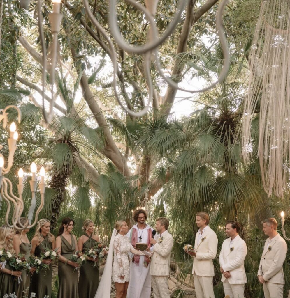 Eine romantische Hochzeitszeremonie im Freien auf Sizilien, mit einem sich an den Händen haltenden Paar, dem Trauredner und den Hochzeitsgästen unter Bäumen und Lichterketten – Heiraten auf Sizilien in seiner bezauberndsten Form.