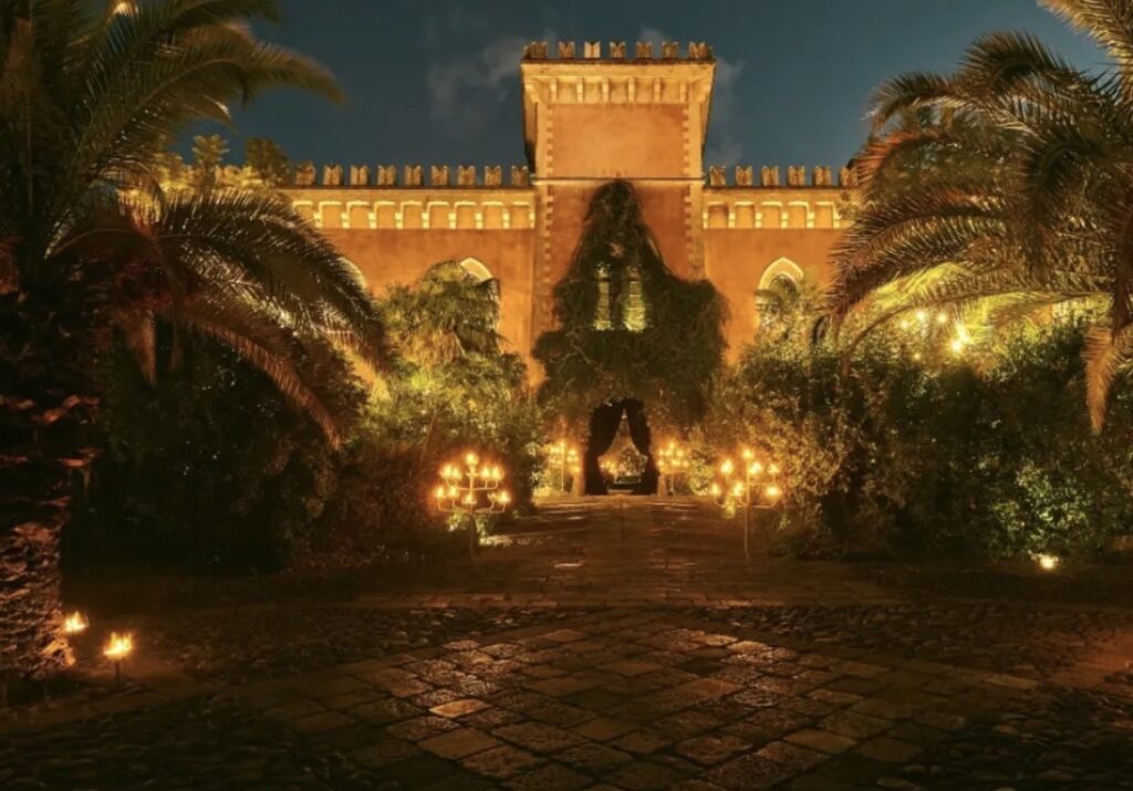 Ein beleuchtetes historisches Gebäude mit Palmen und Kerzen, die nachts einen Gartenweg erhellen – eine bezaubernde Kulisse für „Heiraten auf Sizilien“.