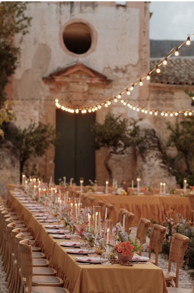 Ein langer Tisch im Freien, gedeckt für ein Abendessen mit Kerzen und Blumen, unter Lichterketten neben einem alten, rustikalen Gebäude – perfekt für eine Hochzeit auf Sizilien.