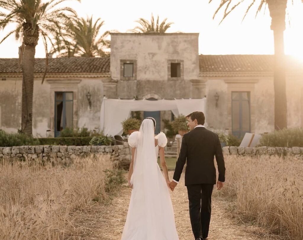 Ein Brautpaar, das sich an den Händen hält, schreitet bei Sonnenuntergang auf ein rustikales Gebäude zu, umgeben von hohem Gras und Palmen – eine bezaubernde Szene, perfekt für eine Hochzeit auf Sizilien.