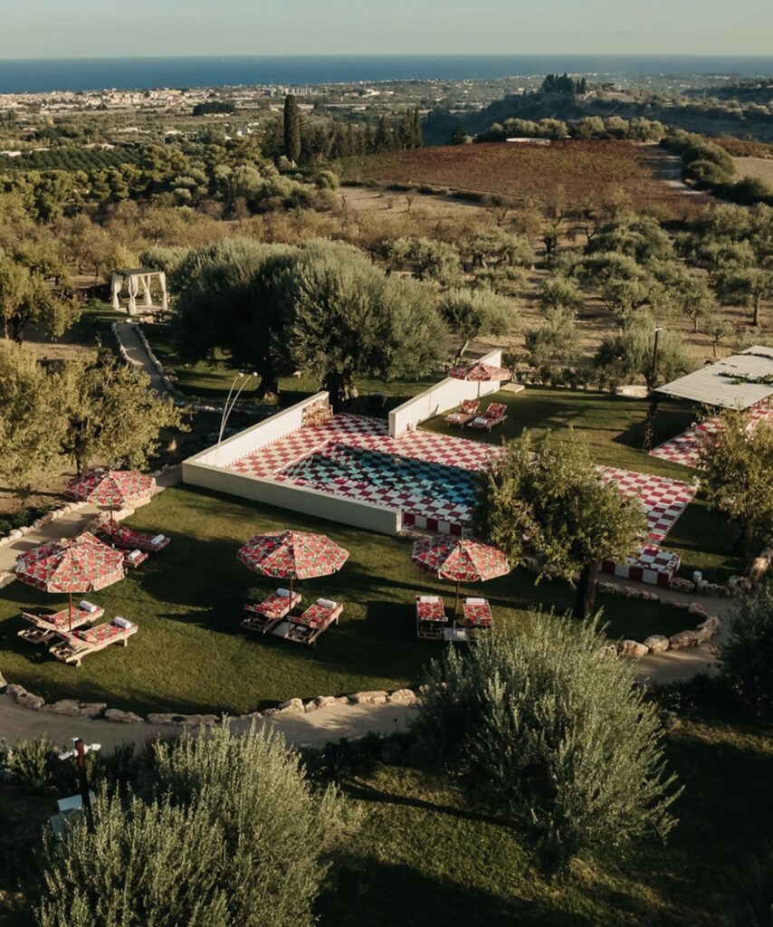Luftaufnahme eines Gartens mit rot karierten Sonnenschirmen, Liegestühlen und einem gemusterten Pool, umgeben von Bäumen – eine idyllische Kulisse für „Heiraten auf Sizilien“.