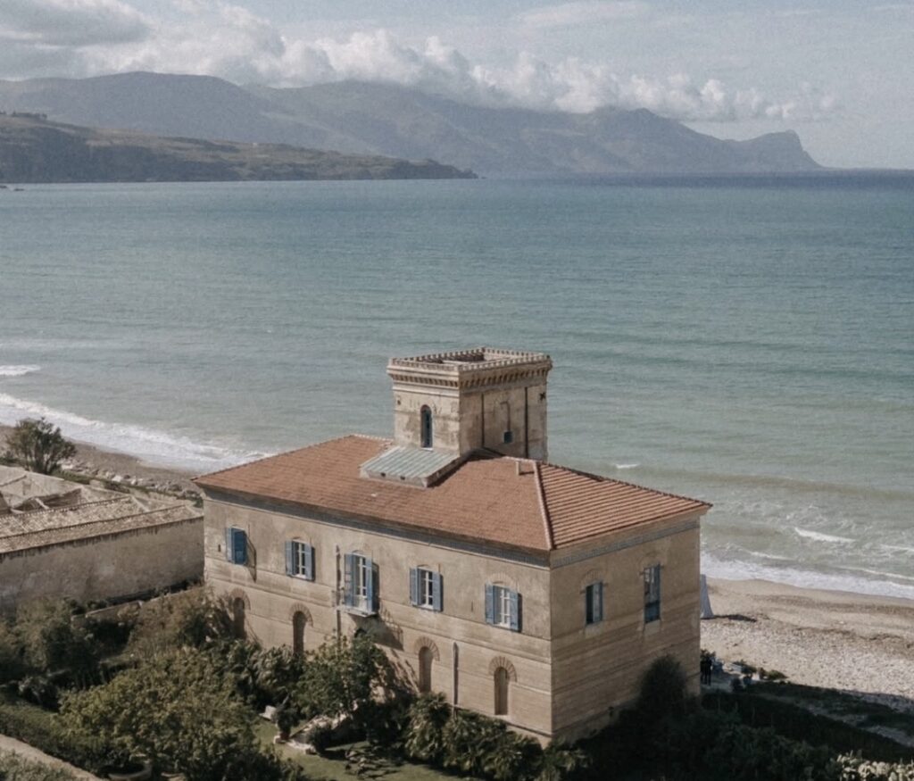 Eine beigefarbene Villa mit rotem Dach, ideal als Hochzeitslocation auf Sizilien, liegt direkt am Meer, mit den Bergen im Hintergrund.