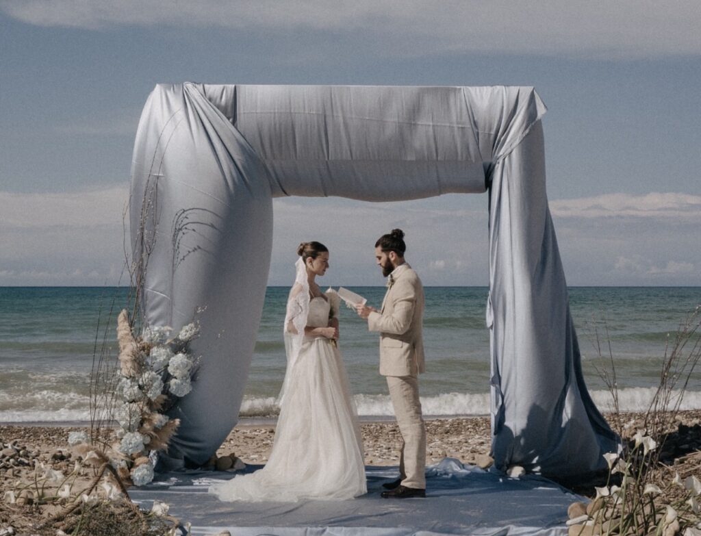 Ein Brautpaar gibt sich unter einem Stoffbogen am Meer das Ja-Wort – einer der beliebtesten Hochzeitsorte auf Sizilien.
