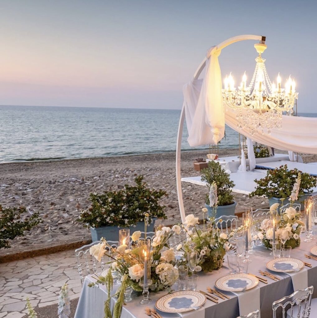 Ein eleganter Tisch am Meer, gedeckt für ein Abendessen mit Blumen, Kerzen und einem Kronleuchter bei Sonnenuntergang – perfekt für Hochzeitslocations auf Sizilien.