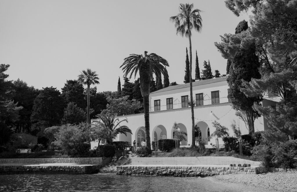 Eine weiße Villa mit Bögen und Palmen direkt am Wasser, umgeben von üppigem Grün, in Schwarz-Weiß – die Villa Dalmacija ist der Inbegriff von Eleganz für alle, die nach Hochzeitslocations in Kroatien suchen oder vom Heiraten am Meer träumen.