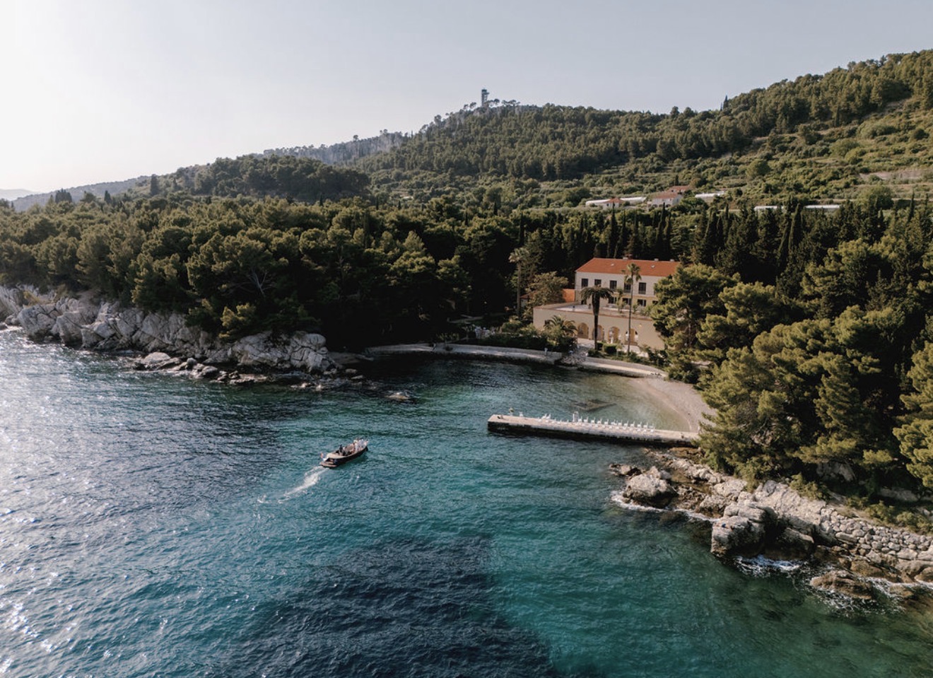 Ein Boot nähert sich einem kleinen Steg bei der Villa Dalmacija an einer bewaldeten Küste mit klarem, blauem Wasser und fängt so den ruhigen Charme ein, der diesen Ort zu einer der begehrtesten Hochzeitslocations in Kroatien für alle macht, die davon träumen, am Meer zu heiraten.