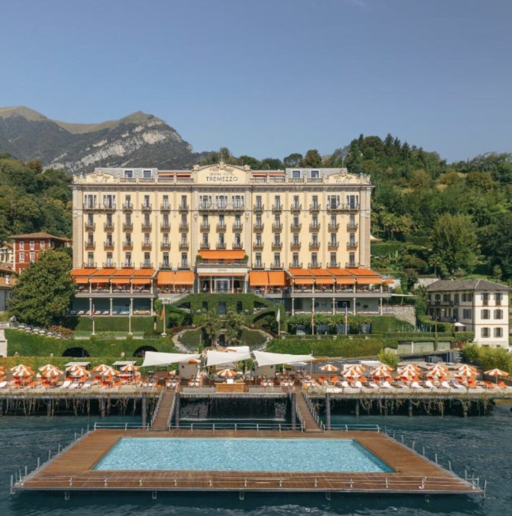 Ein Grand Hotel mit orangefarbenen Markisen, Pool und Sonnenliegen am Seeufer, mit Bergen und grüner Natur im Hintergrund – perfekt für eine luxuriöse Hochzeit im Ausland. Diese atemberaubende Location ist einer der begehrtesten Orte für eine romantische Hochzeit am Comer See.