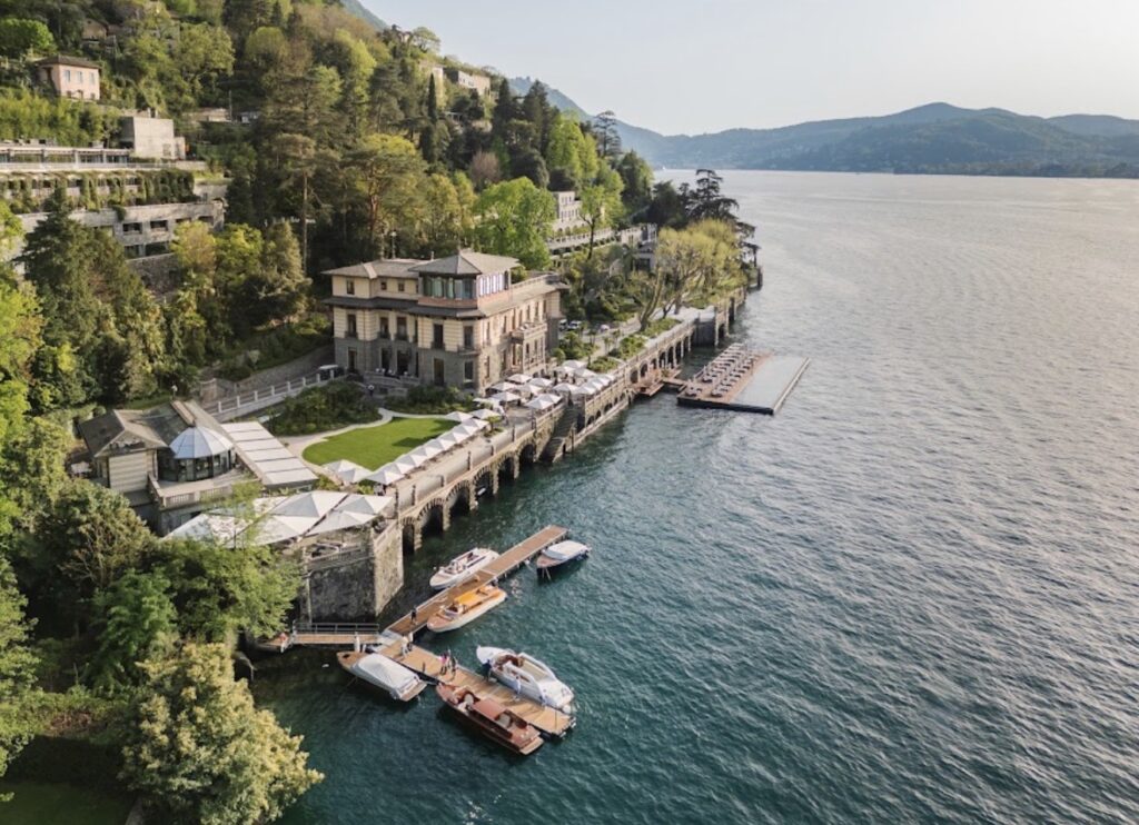 Am Ufer eines Sees, umgeben von Bäumen und Bergen, liegt eine große Villa mit Anlegestellen und Booten – eine idyllische Hochzeitslocation, ideal für eine luxuriöse Hochzeit im Ausland oder eine unvergessliche Hochzeit am Comer See.