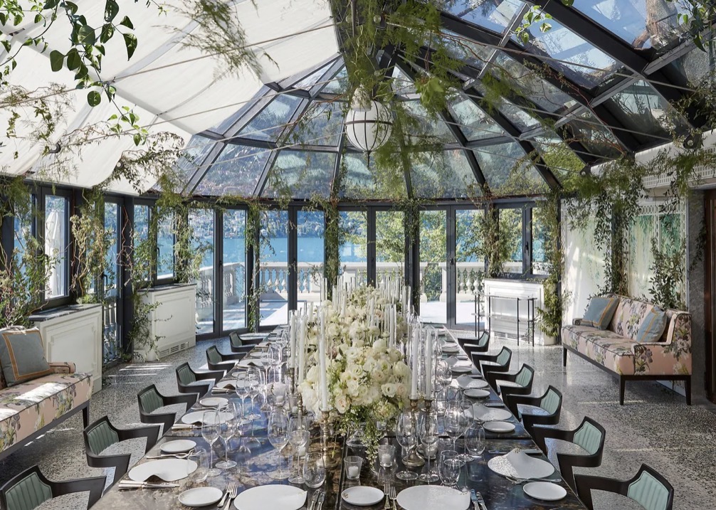 Ein eleganter Speisesaal mit Glasdach, einem langen Tisch, weißen Blumen und Grünpflanzen sowie Blick auf einen blauen See – eine exquisite Kulisse, ideal für Paare, die nach Hochzeitslocations am Comer See suchen oder eine luxuriöse Hochzeit im Ausland planen.