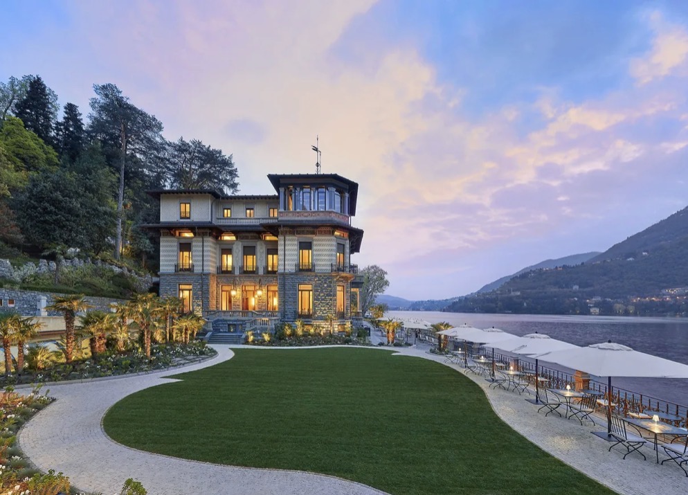 Eine elegante Villa mit leuchtenden Fenstern am Seeufer bei Sonnenuntergang, umgeben von Gärten und Bergen – eine idyllische Kulisse für eine luxuriöse Hochzeit im Ausland oder eine traumhafte Hochzeit am Comer See. Perfekt für Paare, die nach exklusiven Hochzeitslocations in atemberaubender Natur suchen.