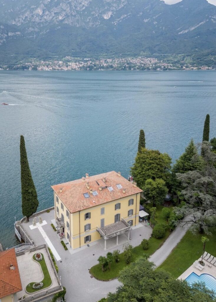 Eine große gelbe Villa mit rotem Dach am Ufer eines Sees, eingebettet inmitten von Bäumen und mit atemberaubendem Blick auf die Berge und ein malerisches Dorf auf der anderen Seite des Sees – eine wahrhaft bezaubernde Kulisse für Hochzeitslocations oder eine luxuriöse Hochzeit im Ausland.