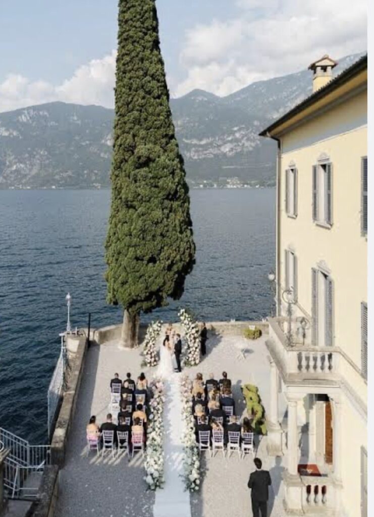 Luxuriöse Hochzeit am Reiseziel: Trauung im Freien am Seeufer, mit sitzenden Gästen und den Bergen im Hintergrund – einer der atemberaubendsten Hochzeitsorte für Ihre unvergessliche Hochzeit am Comer See.