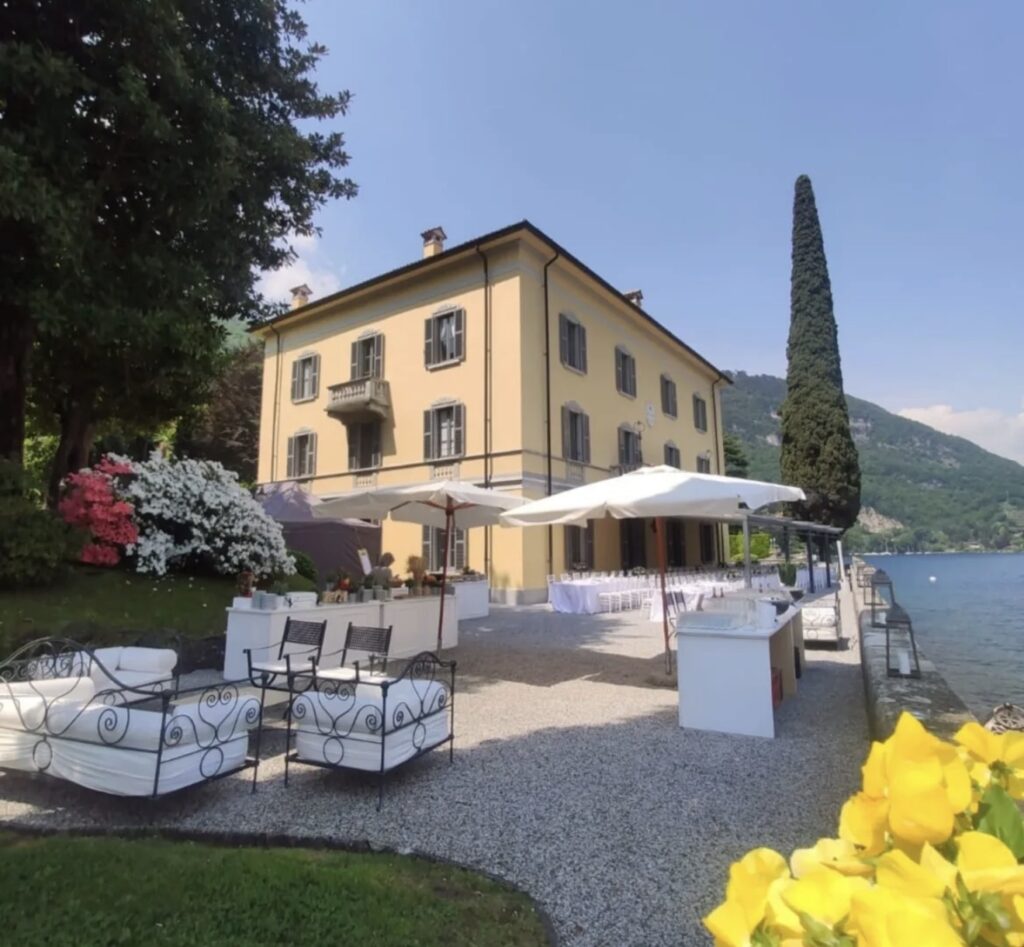 Eine elegante Villa am See mit Sitzgelegenheiten im Freien, weißen Sonnenschirmen, Blumen und den Bergen im Hintergrund an einem sonnigen Tag – ein idealer Ort für eine luxuriöse Hochzeit am Comer See, perfekt für alle, die nach exklusiven Hochzeitslocations suchen.