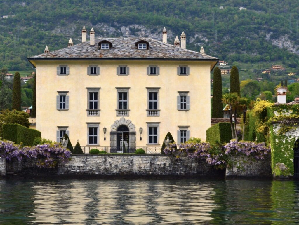 Eine große gelbe Villa mit vielen Fenstern, direkt am See gelegen, umgeben von Gärten und violetten Blumen – dieses atemberaubende Anwesen ist der ideale Ort für eine luxuriöse Hochzeit im Ausland. Es eignet sich perfekt für eine Hochzeit am Comer See und zählt zu den schönsten Hochzeitslocations für Ihren besonderen Tag.