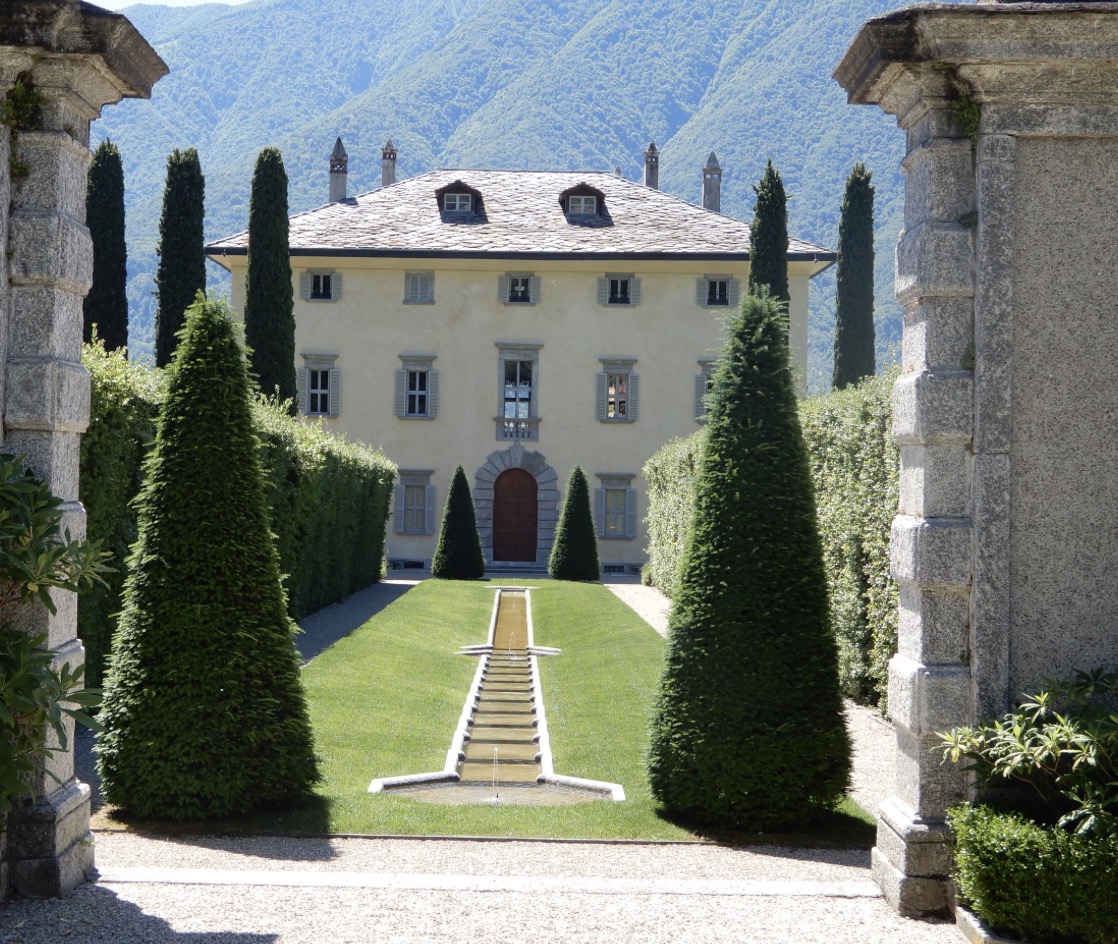 Ein gepflegter Garten mit symmetrischen Hecken, der zu einer großen Villa führt, eingerahmt von Steinsäulen und den Bergen im Hintergrund – eine idyllische Kulisse, perfekt für eine luxuriöse Hochzeit im Ausland oder für alle, die nach exquisiten Hochzeitslocations für eine unvergessliche Hochzeit am Comer See suchen.