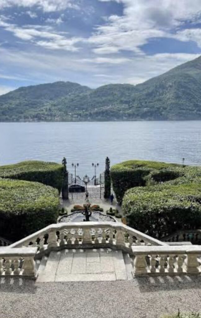 Steinstufen führen zu gepflegten Hecken, einem Tor und einem See mit Hügeln im Hintergrund unter bewölktem Himmel – eine bezaubernde Kulisse, die sich perfekt für eine Hochzeit am Comer See eignet und diesen Ort zu einer der begehrtesten Hochzeitslocations für eine luxuriöse Hochzeit im Ausland macht.