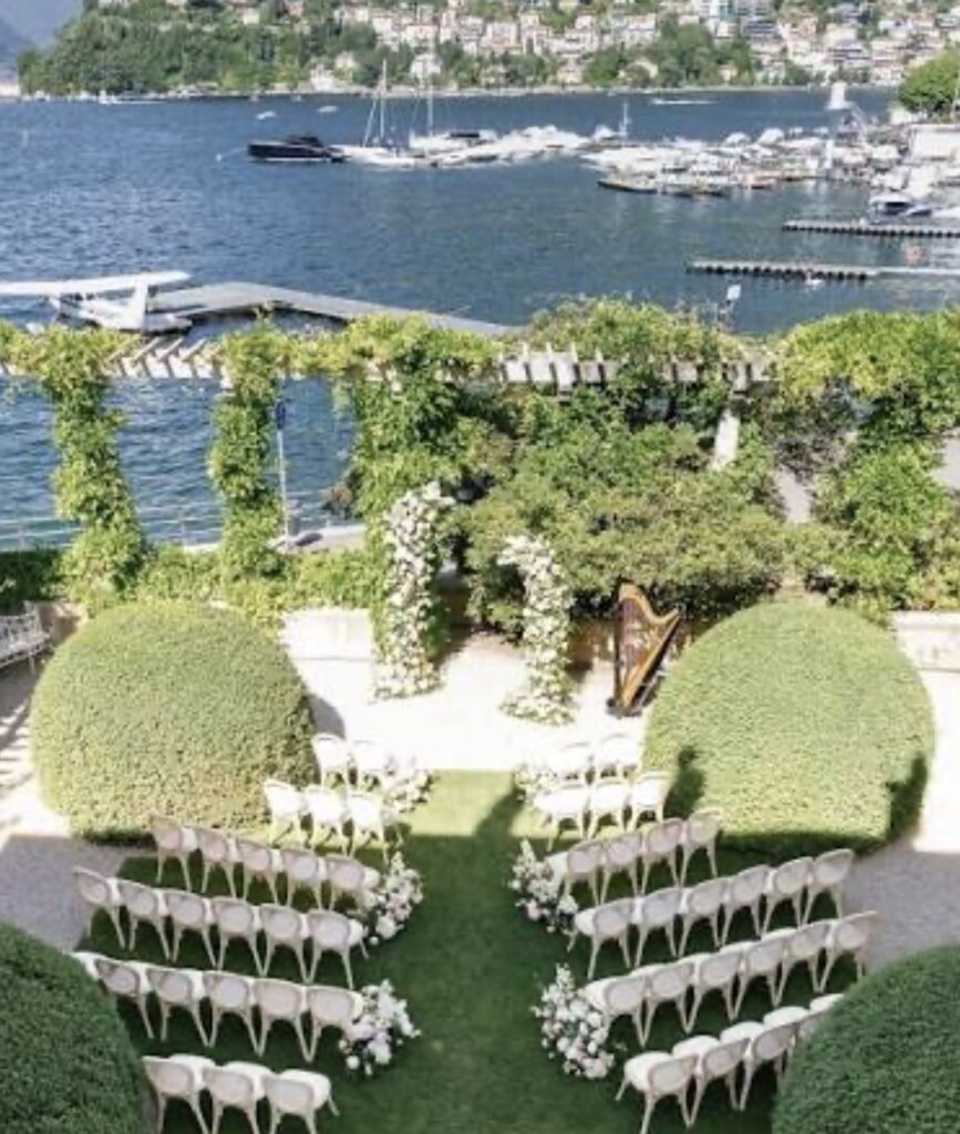 Luxuriöse Hochzeit am Reiseziel: Eine Hochzeitskulisse im Freien am Seeufer mit weißen Stühlen, einem mit Blumen geschmückten Gang und einer Harfe, mit Blick auf Boote und Hügel – eine exquisite Wahl für Ihre Hochzeit am Comer See.