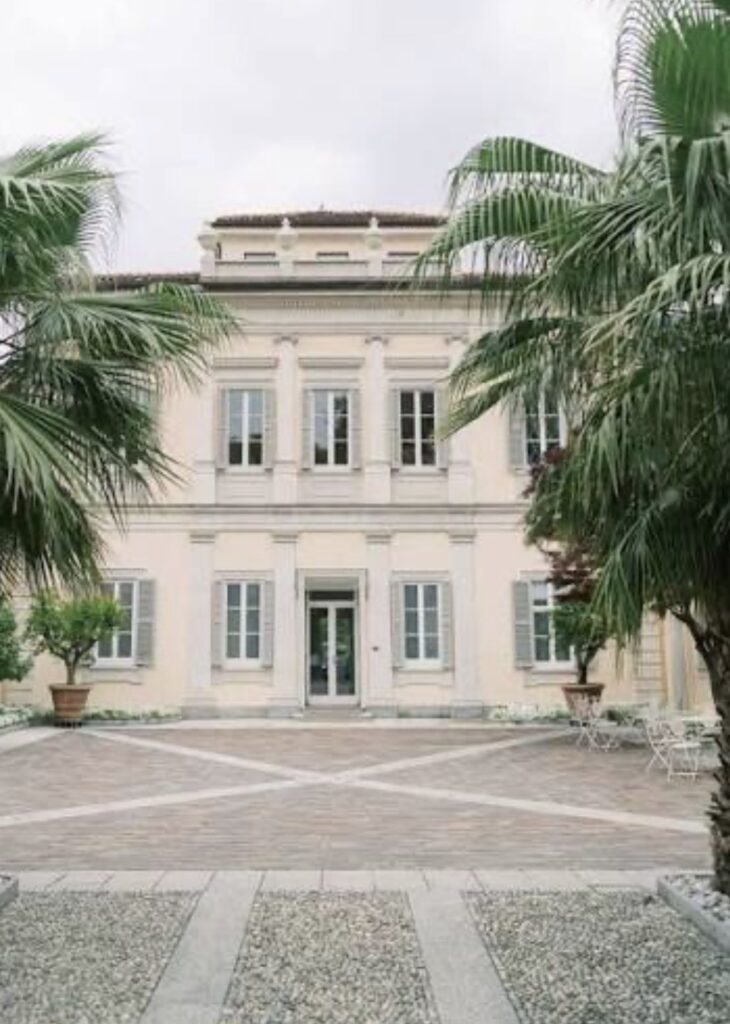 Ein cremefarbenes, zweistöckiges Gebäude mit Palmen, Fenstern und einem steinernen Innenhof davor – eine exquisite Kulisse, die sich perfekt für eine luxuriöse Hochzeit im Ausland eignet oder als einer der begehrtesten Hochzeitsorte für Ihre Hochzeit am Comer See.