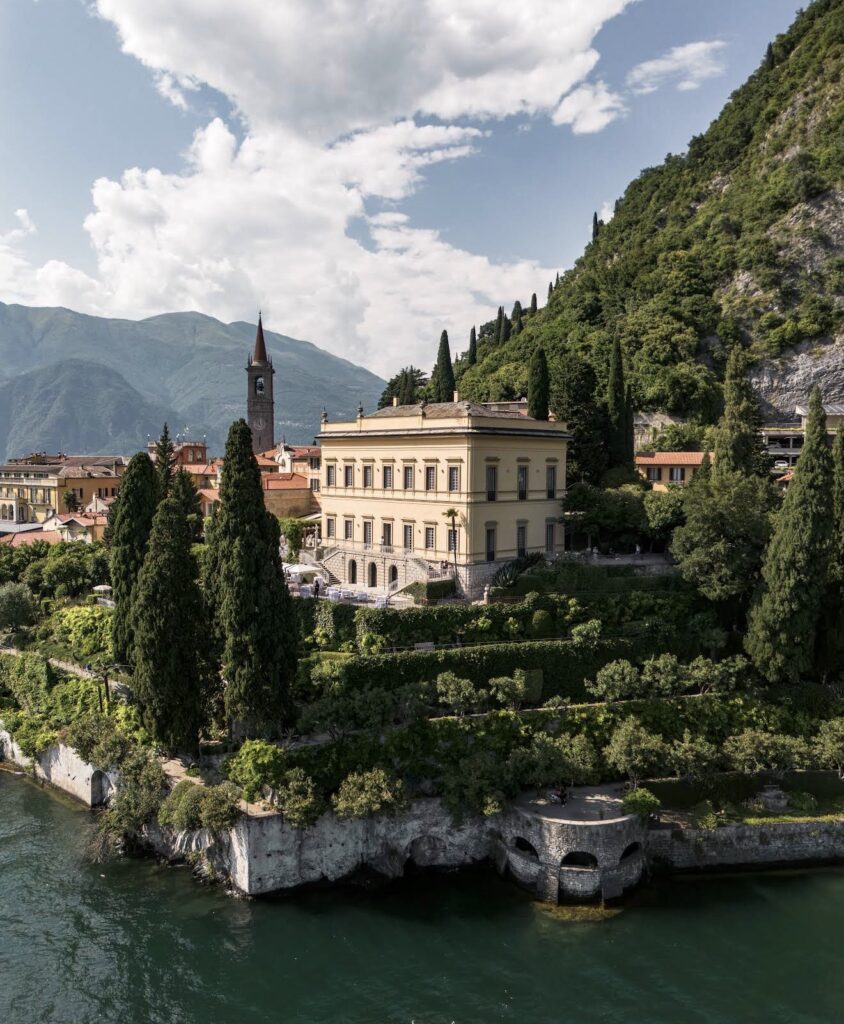 Eine prächtige Villa, umgeben von hohen Bäumen und viel Grün, direkt an einem See und inmitten von Hügeln unter einem teilweise bewölkten Himmel gelegen – dieses Anwesen zählt zu den begehrtesten Hochzeitslocations für eine luxuriöse Hochzeit am Comer See.