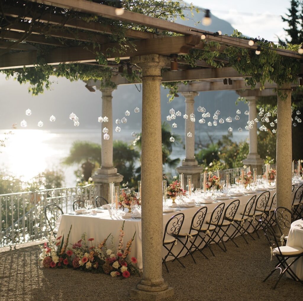 Ein im Freien aufgestelltes Banketttisch-Set für eine Veranstaltung am See, mit Blumendekorationen und hängenden Glaskugeln – eine bezaubernde Kulisse, die sich perfekt für eine luxuriöse Hochzeit an einem besonderen Ort eignet. Diese atemberaubende Kulisse verkörpert den Charme exklusiver Hochzeitslocations und ist ideal für Ihre unvergessliche Hochzeit am Comer See.
