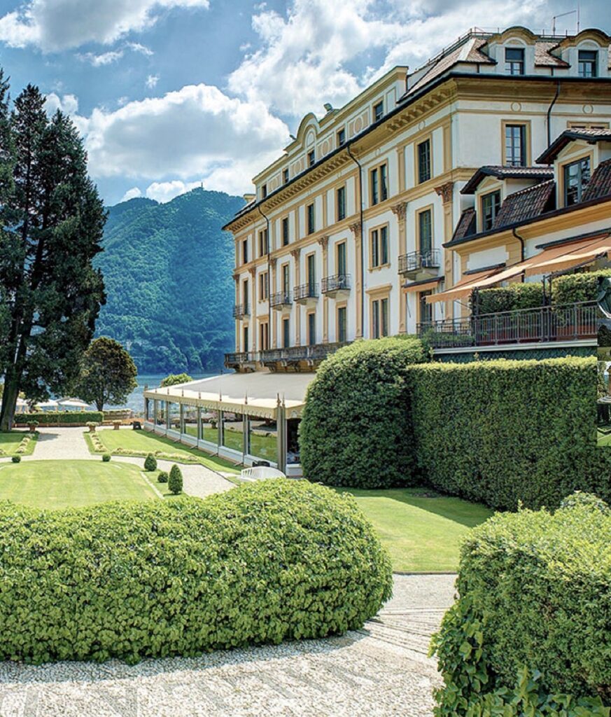 Ein elegantes Hotel mit gepflegten Gärten, Hecken und Blick auf die Berge unter einem blauen Himmel mit vereinzelten Wolken – eine idyllische Kulisse am Ufer des Comer Sees, perfekt für alle, die auf der Suche nach Hochzeitslocations oder einer luxuriösen Hochzeit im Ausland sind.